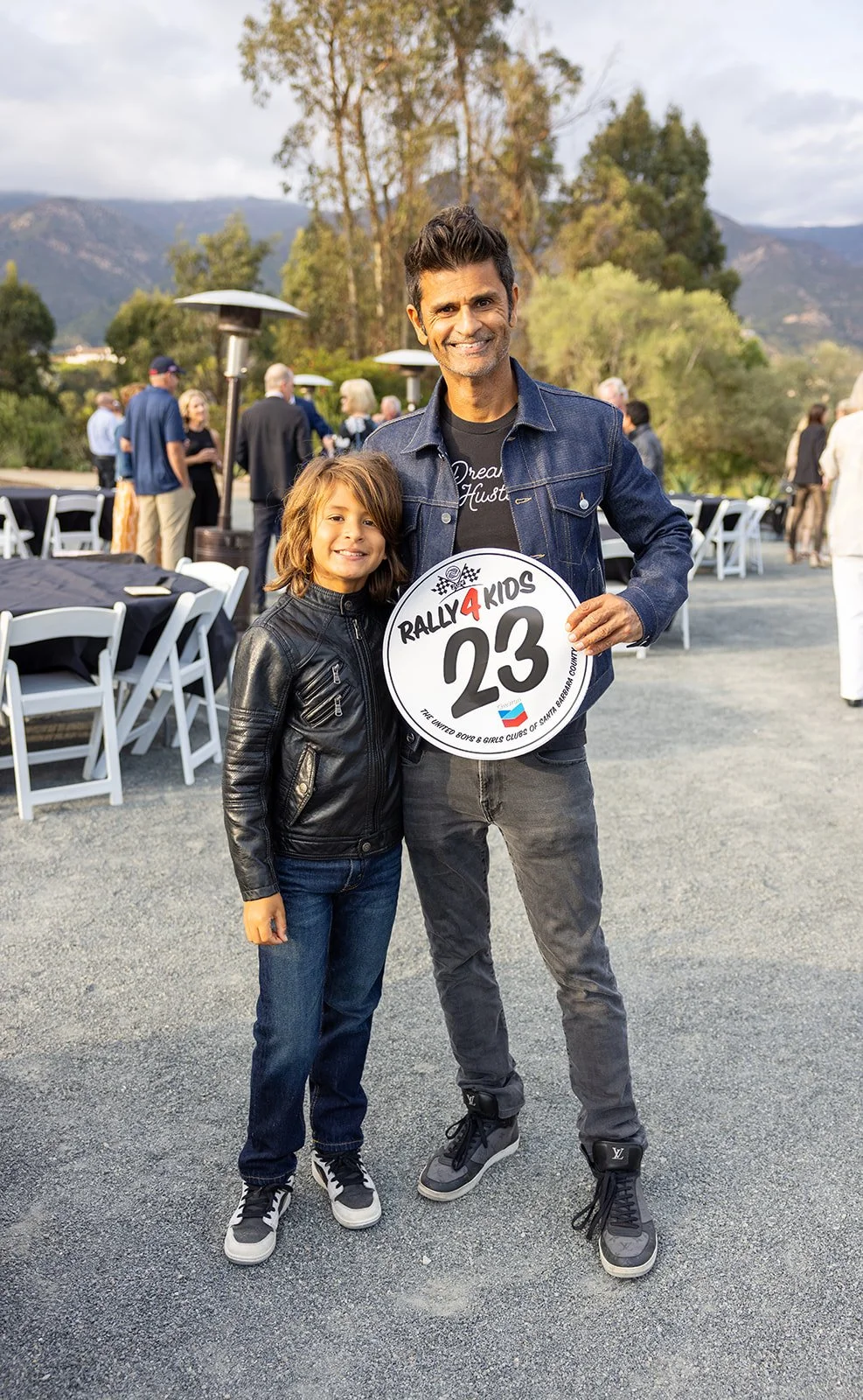 A man and a young girl smiling and standing outdoors at a gathering, the man holding a circular sign that says 'Rally4Kids 23'. There are tables with chairs and other people in the background, with trees and mountains in the distance.