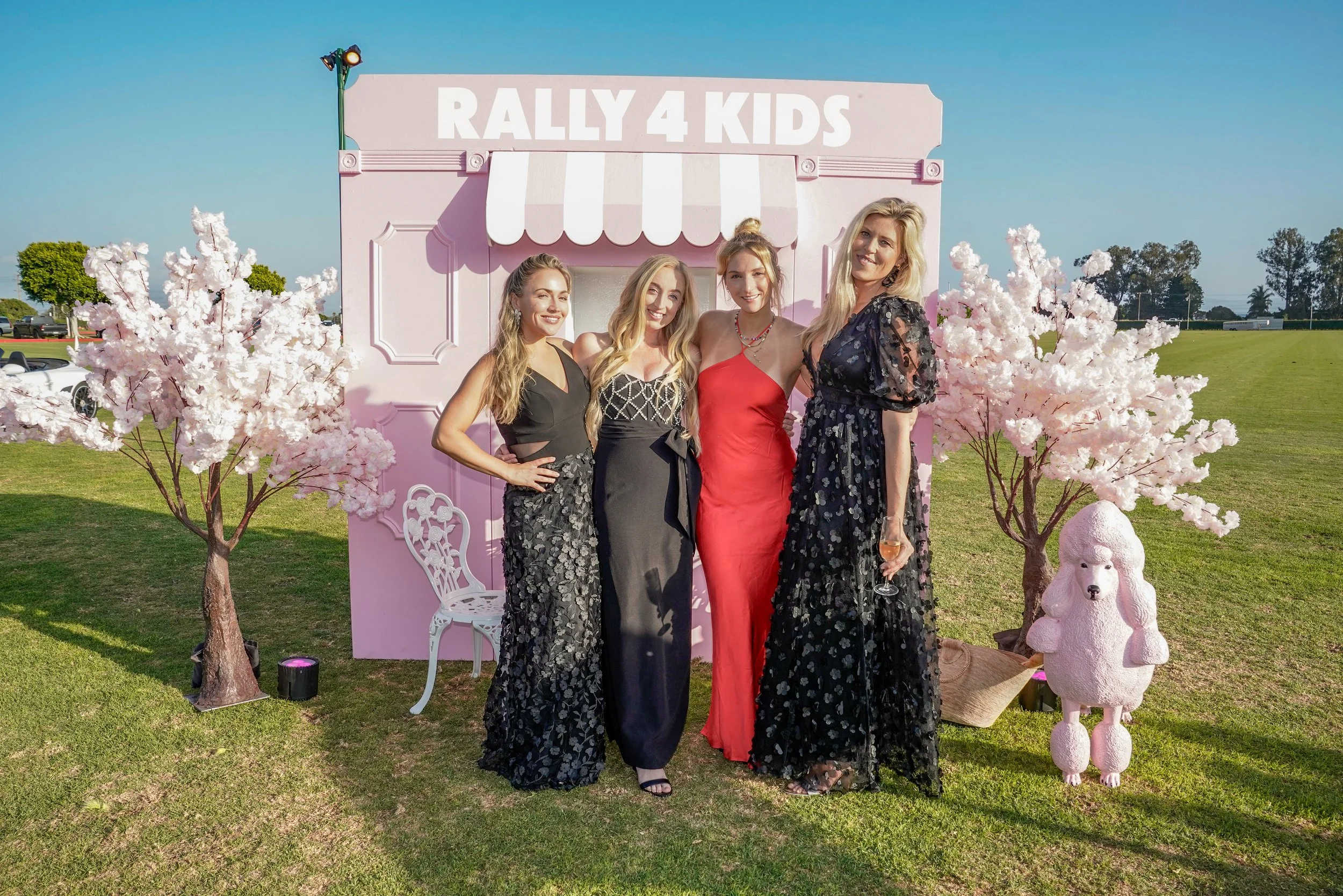 Four women standing in front of a pink backdrop with a sign that reads 'Rally 4 Kids.' The backdrop is decorated with pink cherry blossom trees and plush pink poodle statues. The women are dressed in formal evening gowns, smiling and posing for the p