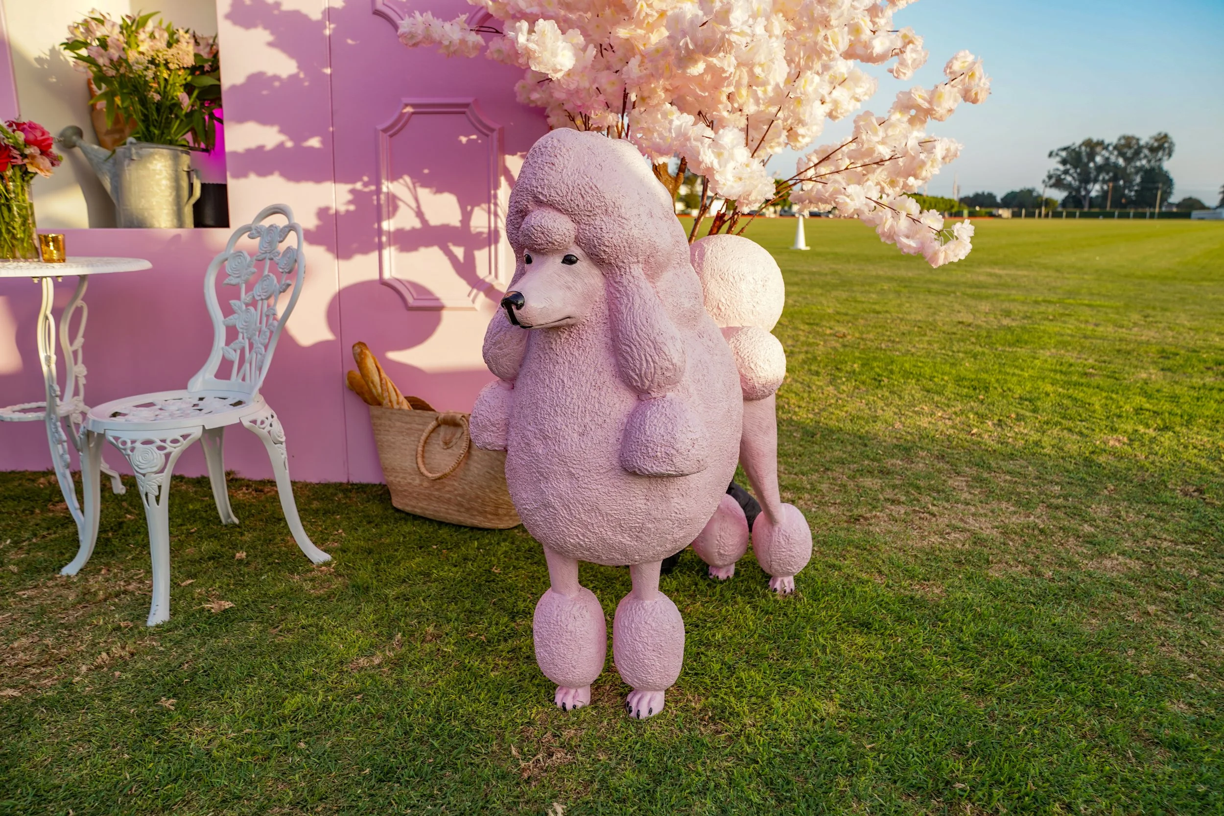 Decorative pink poodle sculpture with pompom details in a garden setting, featuring pink cherry blossom branches, a pink backdrop, and garden furniture.