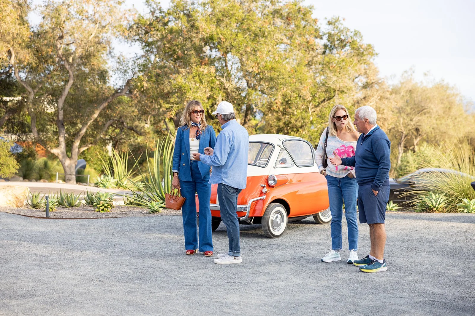 Four people standing outdoors near an orange and white vintage microcar, engaging in conversation. The group includes two women and two men, all dressed casually. The background features trees and shrubbery, suggesting a park or garden setting.