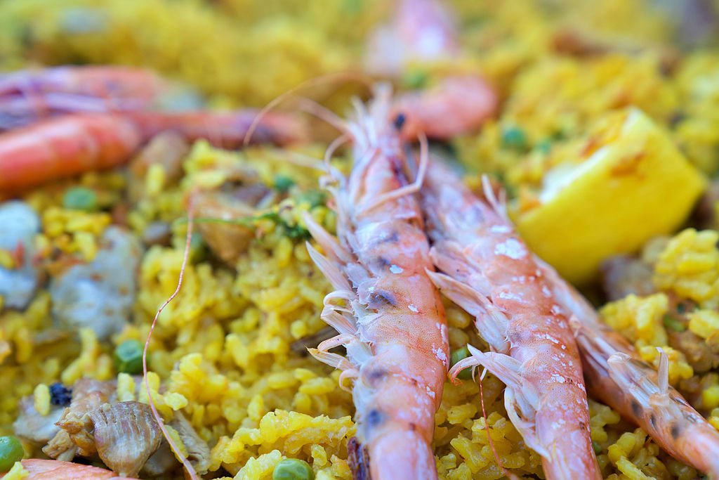 Close-up of a seafood dish with pink shrimp on top of yellow rice with vegetables and lemon wedge.