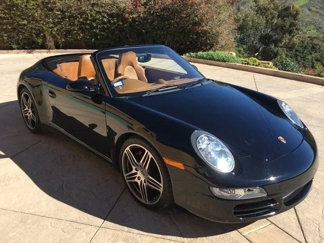 Black Porsche convertible with tan interior parked on a driveway with trees in the background.