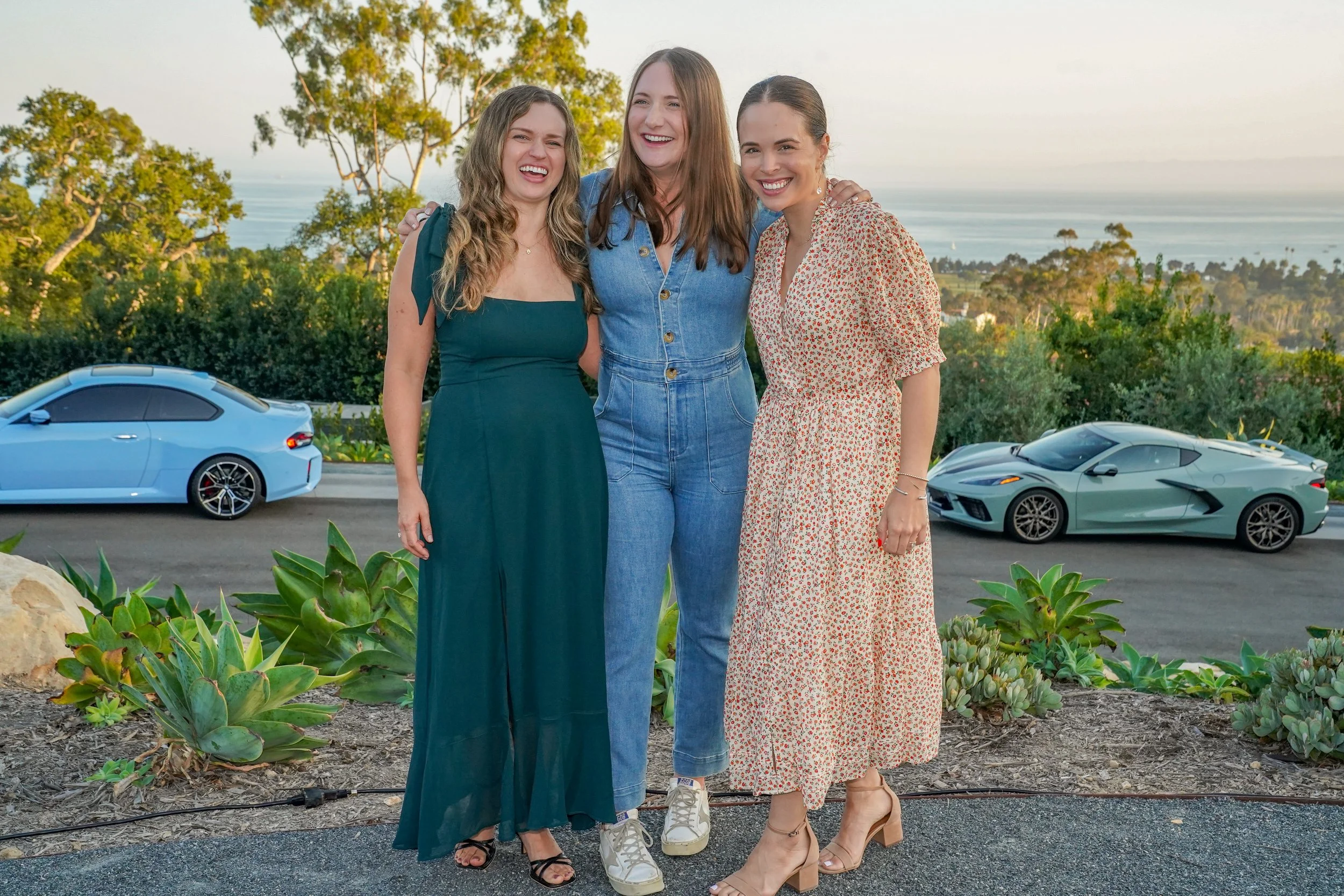 Three women standing outdoors with arms around each other, smiling, with luxury cars and a scenic view of trees, water, and the horizon in the background.