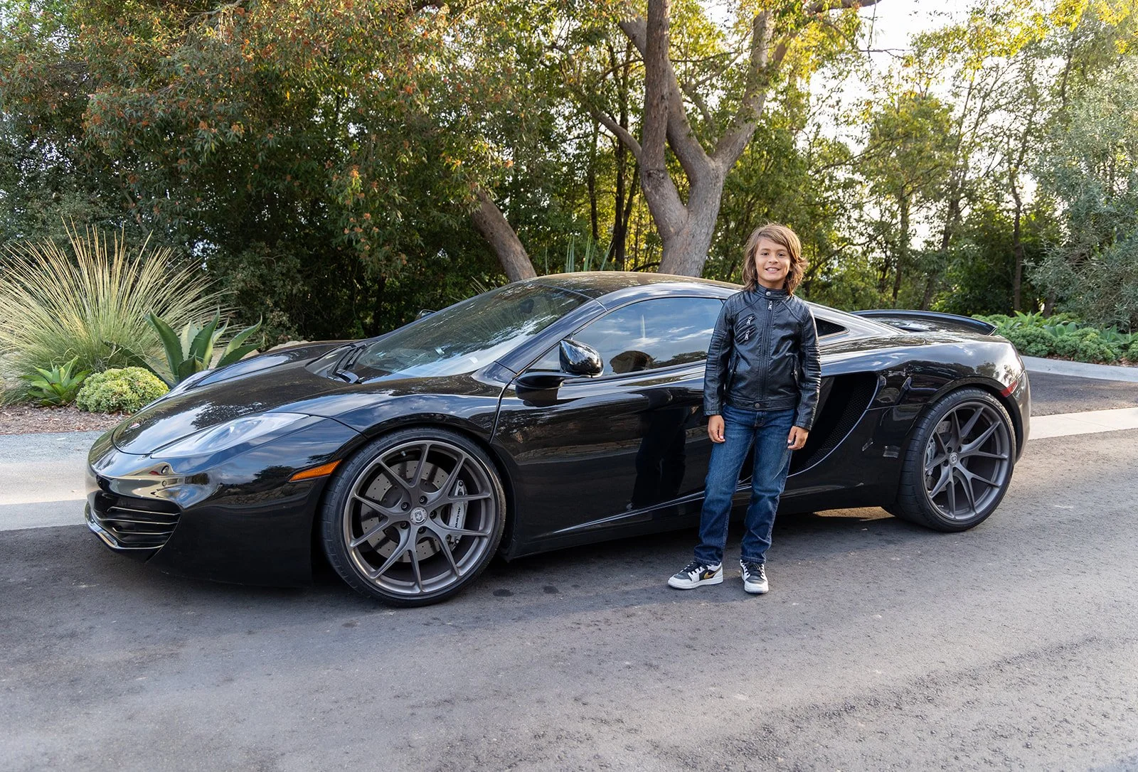 A young boy with long hair smiling, wearing a black leather jacket, jeans, and sneakers, standing next to a black sports car on a street with trees and greenery in the background.