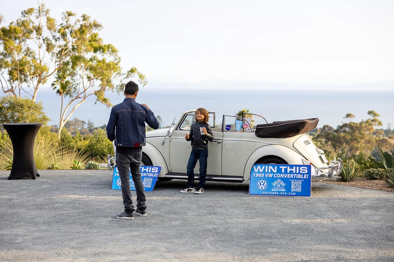 A woman and a man near a white vintage convertible car with signage for a contest to win a 1960 VW convertible, set outdoors with trees and a view of the water in the background.