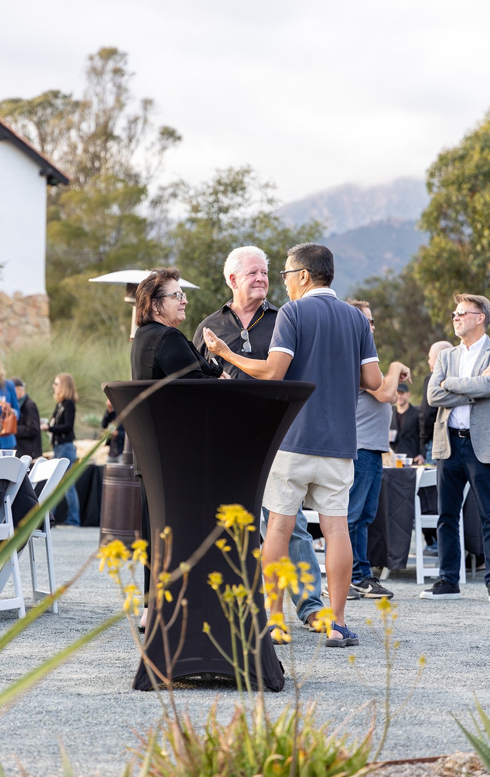 People socializing outdoors at a gathering, with a black high-top table in the foreground, some seated and standing under a partly cloudy sky, surrounded by mountains and trees.