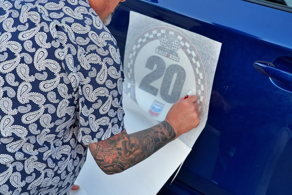 A man with tattoos on his arm is sticking a large sticker or decal on the side of a blue car. The decal features a black and white checkered border, the number 20, and the text 'Rally Kids'.