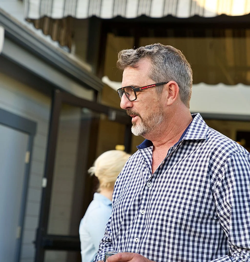 A middle-aged man with glasses, gray hair, and a beard, wearing a blue and white checkered shirt, standing outdoors.