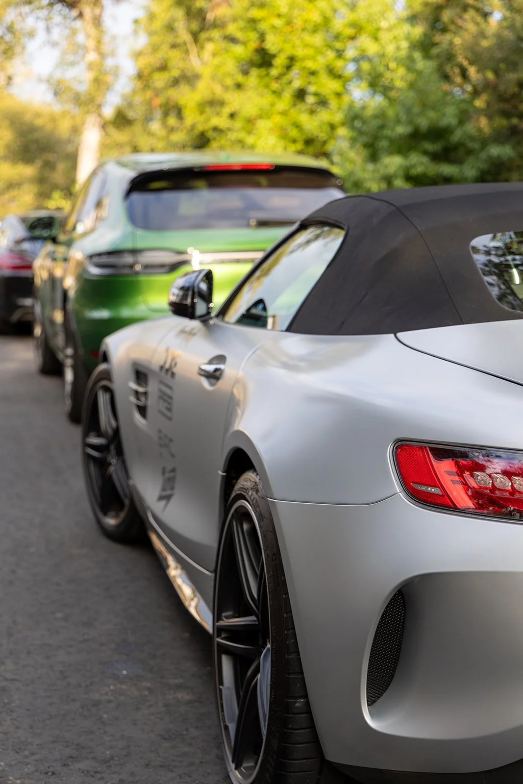Close-up of a silver convertible sports car parked outdoors, with black wheels and a black soft top, in front of a green SUV, during daytime with trees in the background.