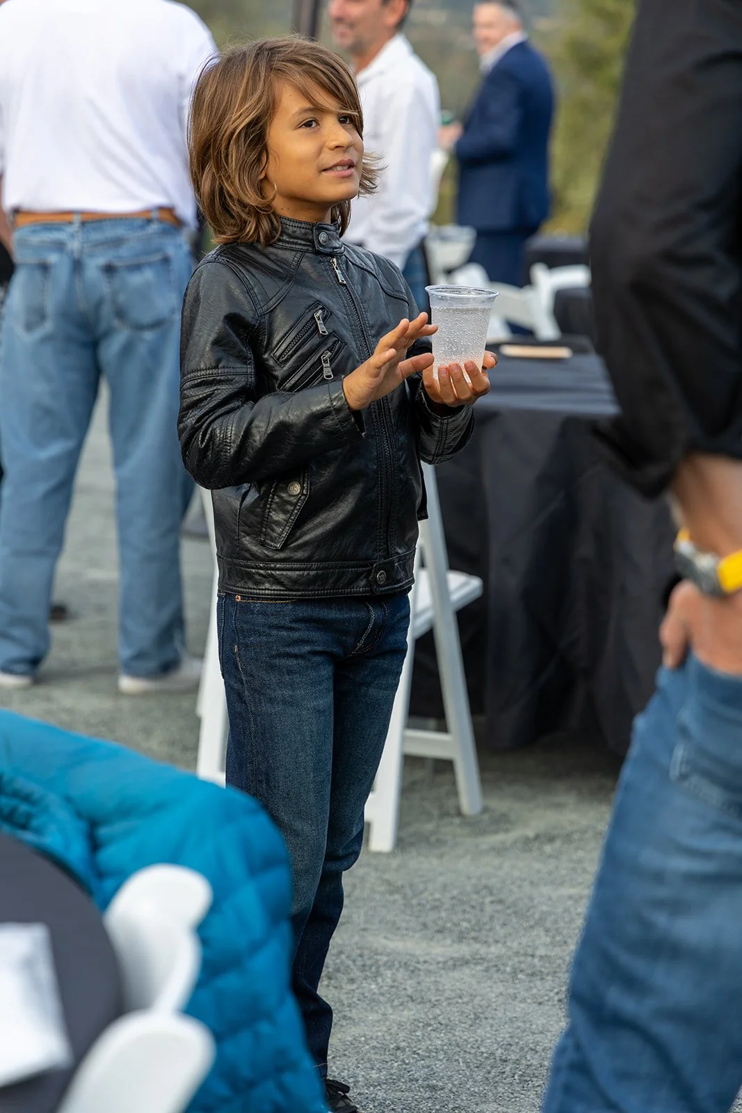 A young boy with brown hair wearing a black leather jacket and jeans holding a clear plastic cup with a bubbly drink at an outdoor gathering.