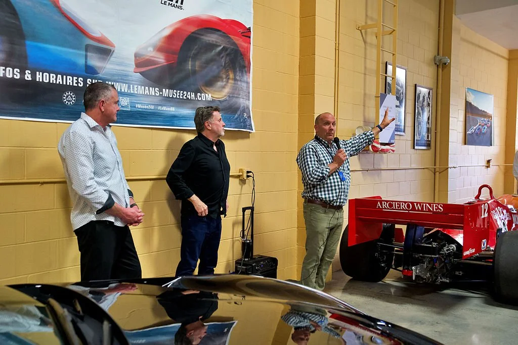 Three men standing beside a red Formula 1 race car and a large banner with a race car graphic. One man is speaking into a microphone, pointing at a picture on the wall. The setting appears to be an indoor racing or motorsport event.