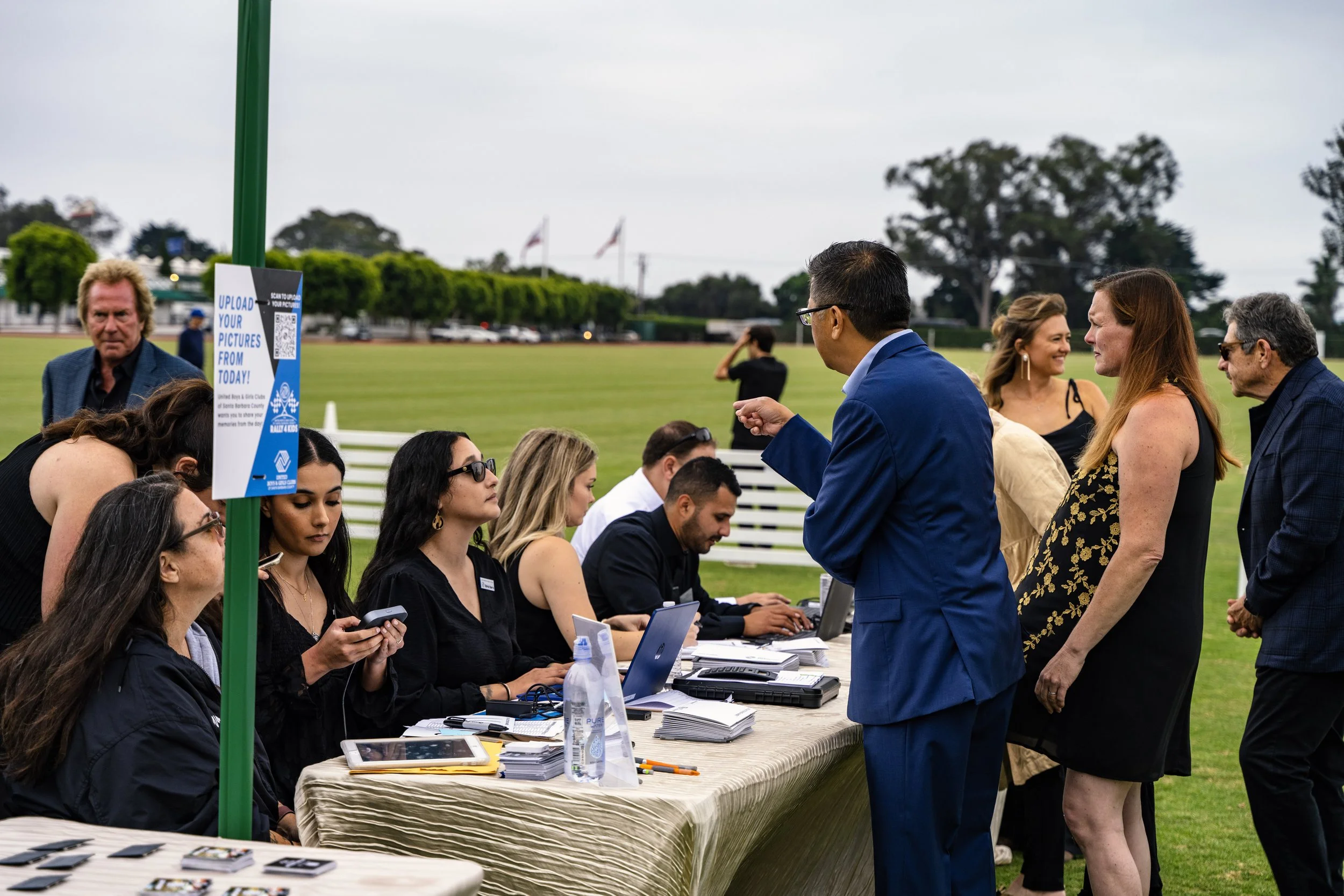 People in line at outdoor registration or check-in table with registration staff, on a grassy field with trees and flags in the background.