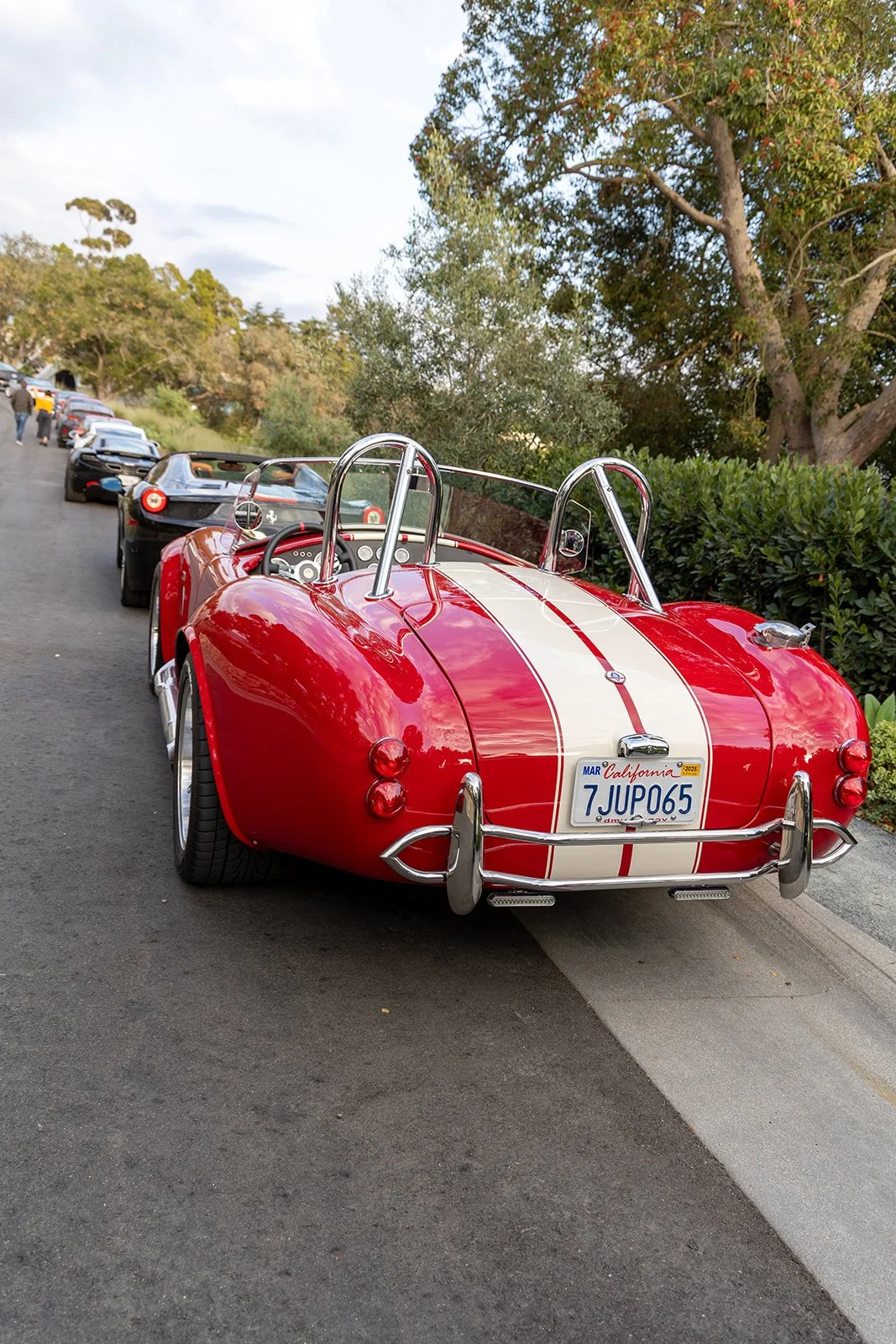 Red vintage sports car with white racing stripes parked on a street in front of trees and hedges, with other cars lined up behind it.