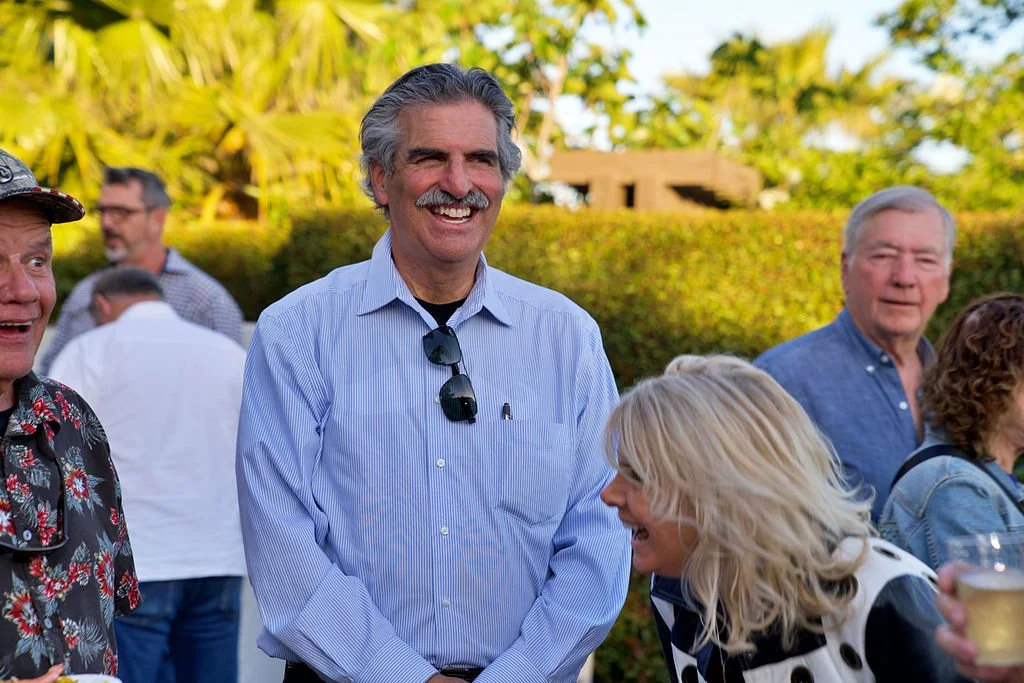 Group of people outdoors enjoying a social gathering, smiling and laughing, with greenery and trees in the background.