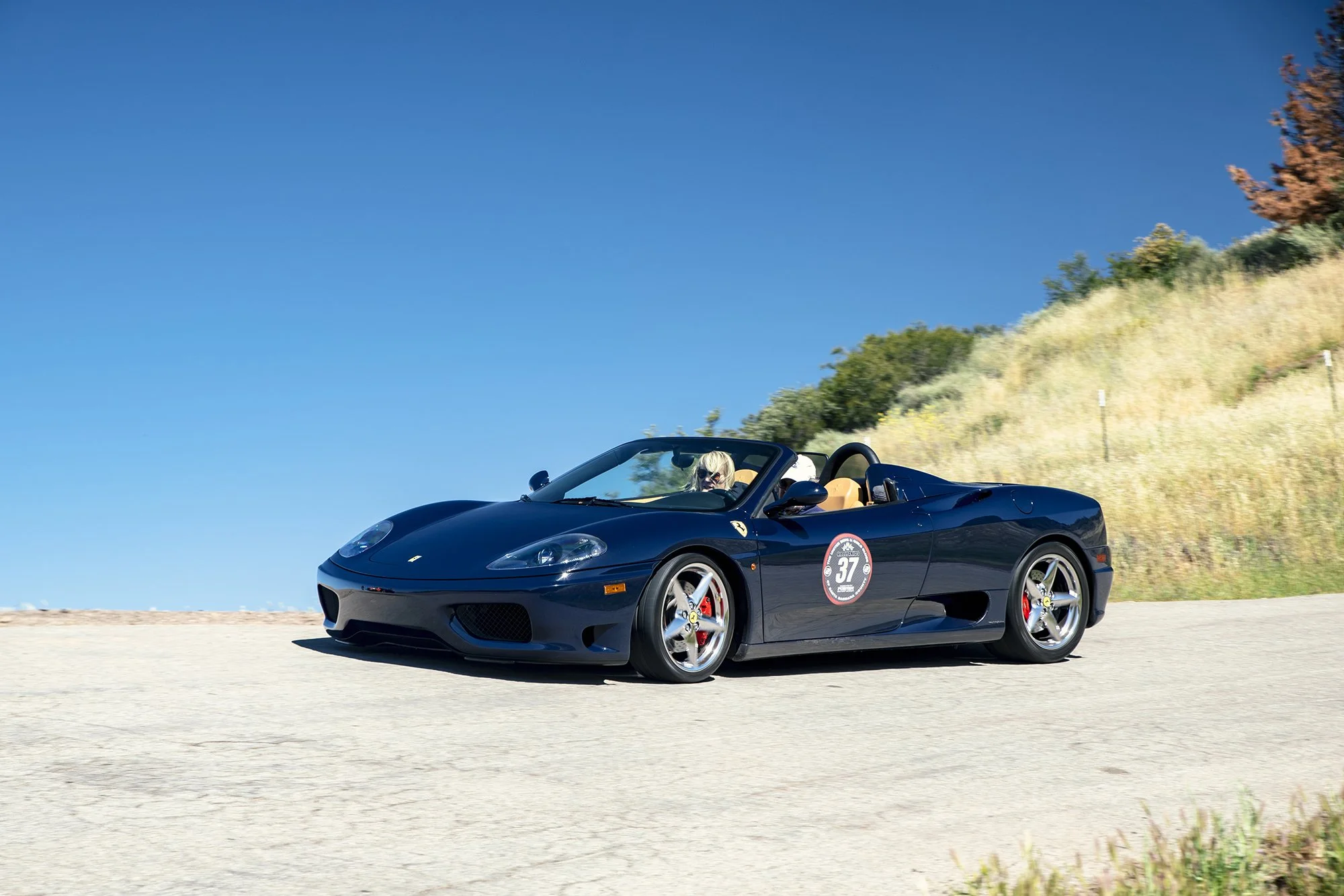 A dark blue Ferrari convertible sports car with beige interior, driven by a woman with blonde hair and sunglasses, on a clear day with blue sky and grassy hillside in the background.