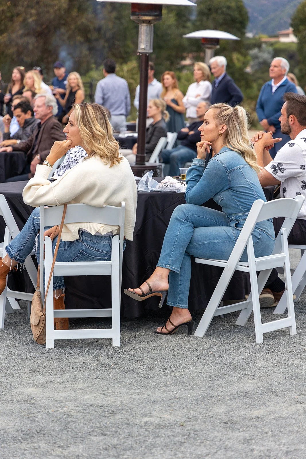 People attending an outdoor event, sitting at chairs and tables, with trees and mountains in the background, some standing and some sitting, dressed casually.