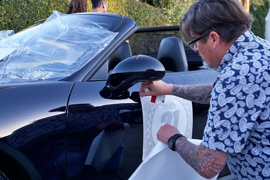 A man with tattoos and glasses applies a race number decal to the side of a black convertible car.