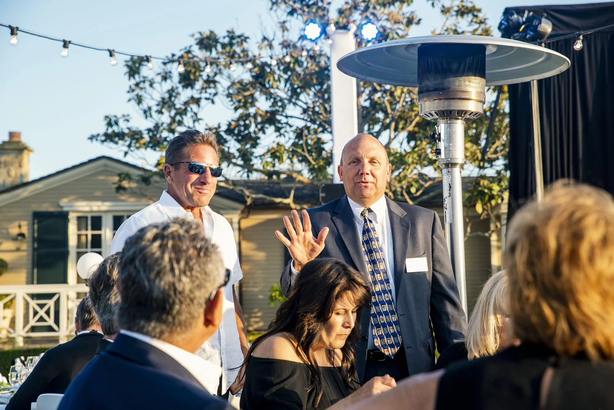 Man in suit and tie speaking at outdoor gathering with seated guests, another man in sunglasses standing beside him, patio heater in background, houses and trees in the background.