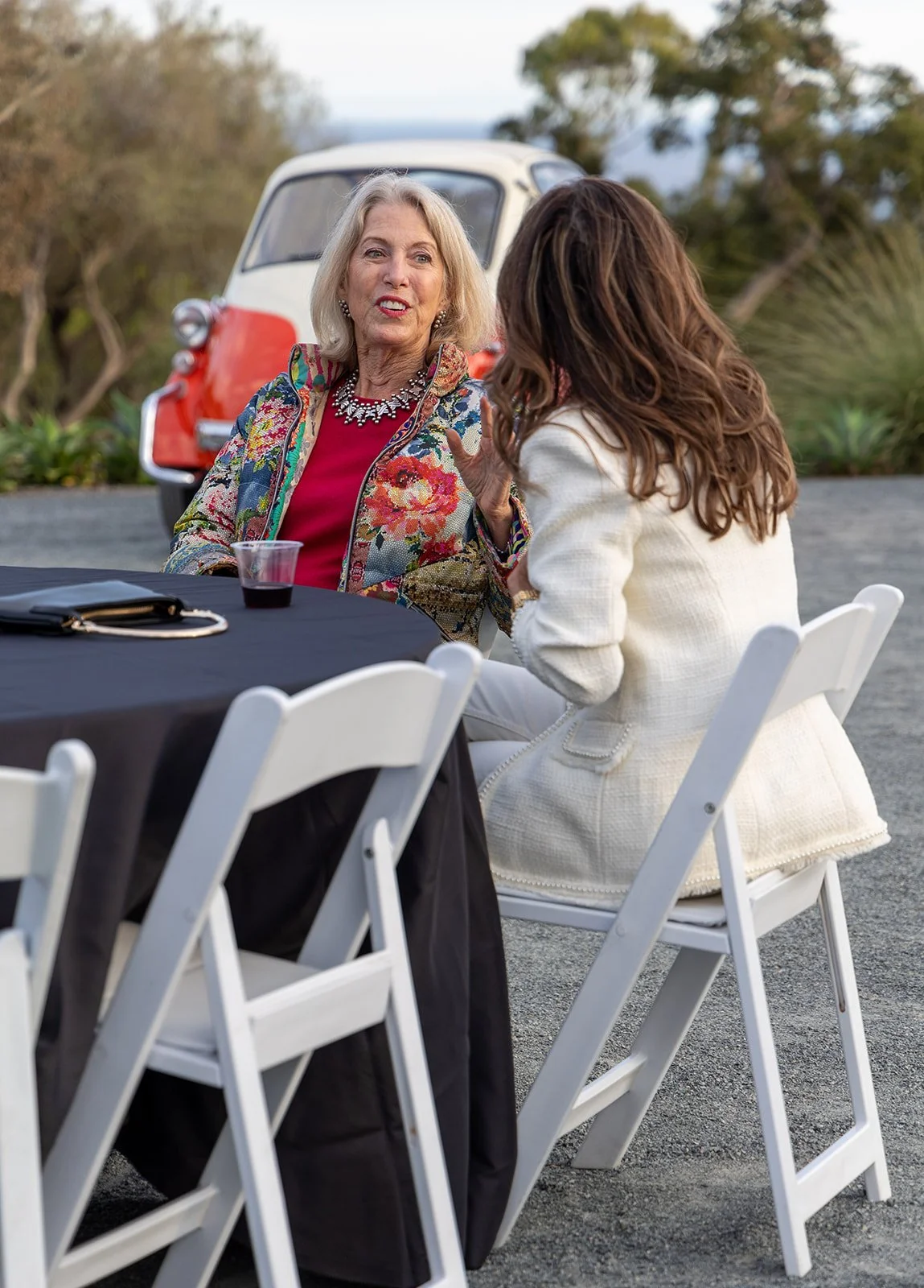 Two women engaged in conversation at an outdoor event, sitting at a black table with white folding chairs; a vintage red and white vehicle and trees are in the background.