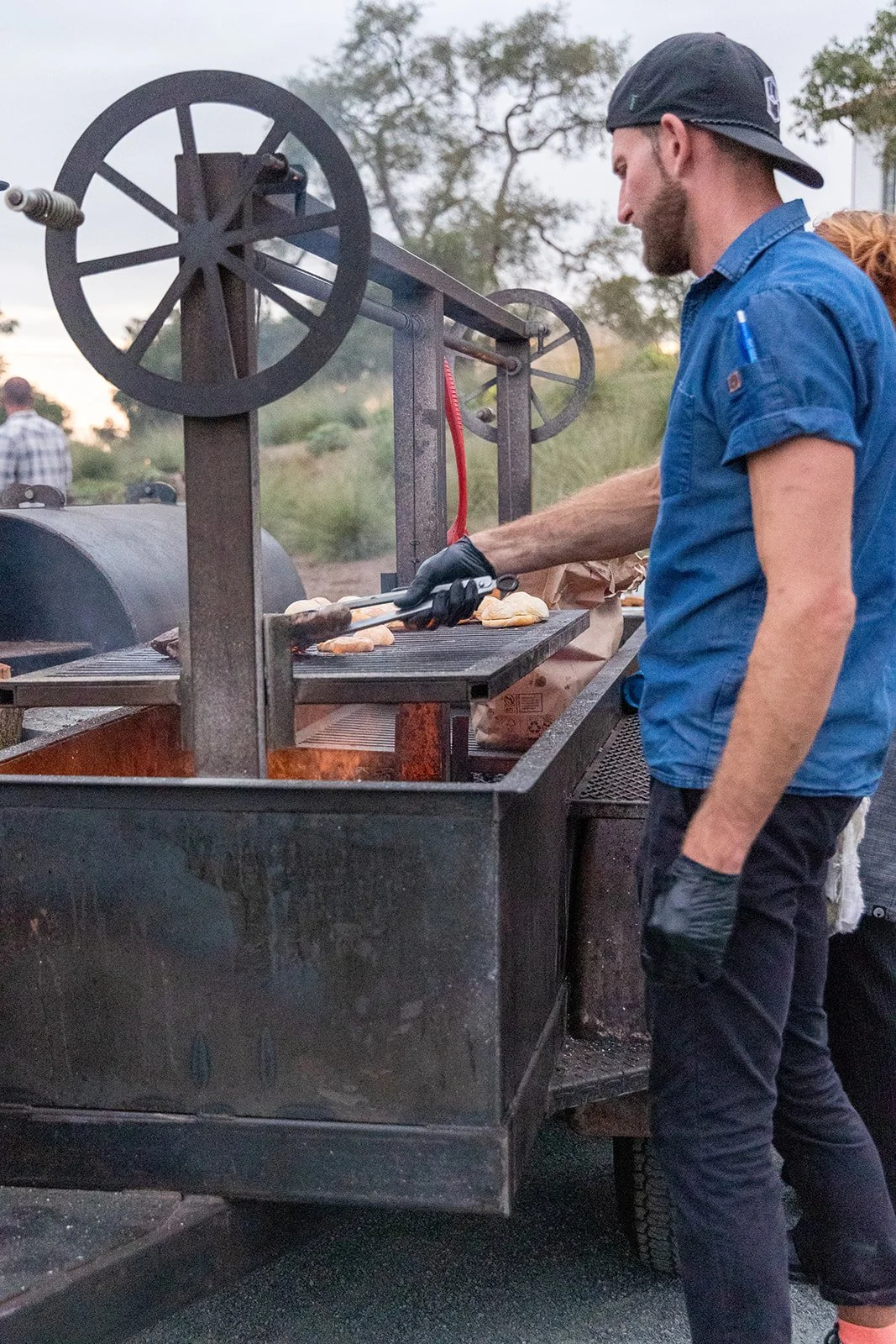 A man grilling chicken on a barbecue smoker outdoor during sunset, with another person nearby and trees in the background.