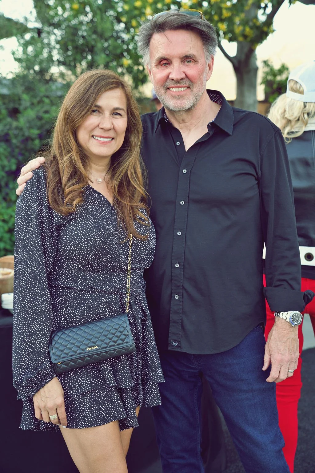 A woman and a man posing outdoors, smiling. The woman has long, wavy brown hair and is wearing a black polka dot dress with long sleeves, a Prada purse, and is accessorized with jewelry. The man has gray hair and a beard, wearing a black button-up sh