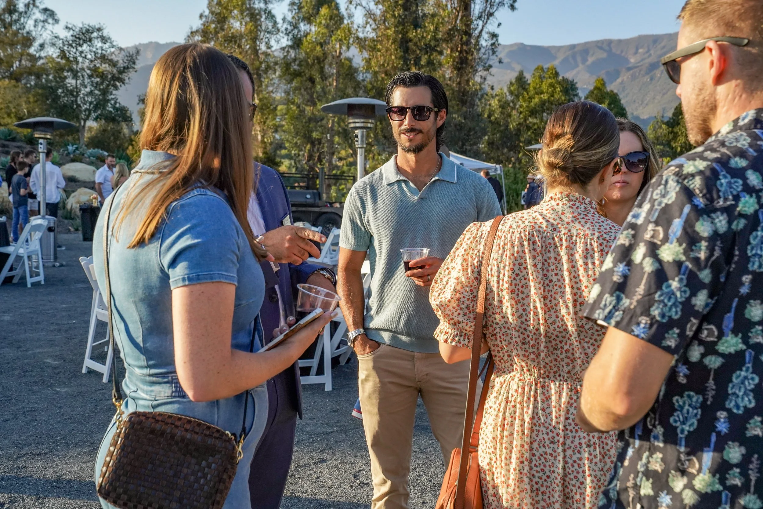A group of people socializing outdoors at a gathering or party, with mountains and trees in the background during sunset.