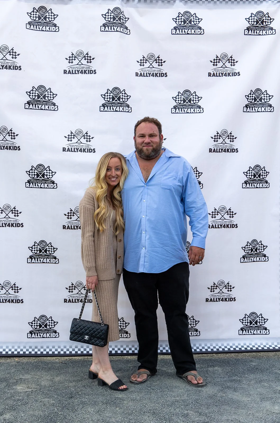 A woman and a man posing together at an event with a backdrop that says 'Rally4Kids' and features crossed checkered flags and a tennis ball logo.