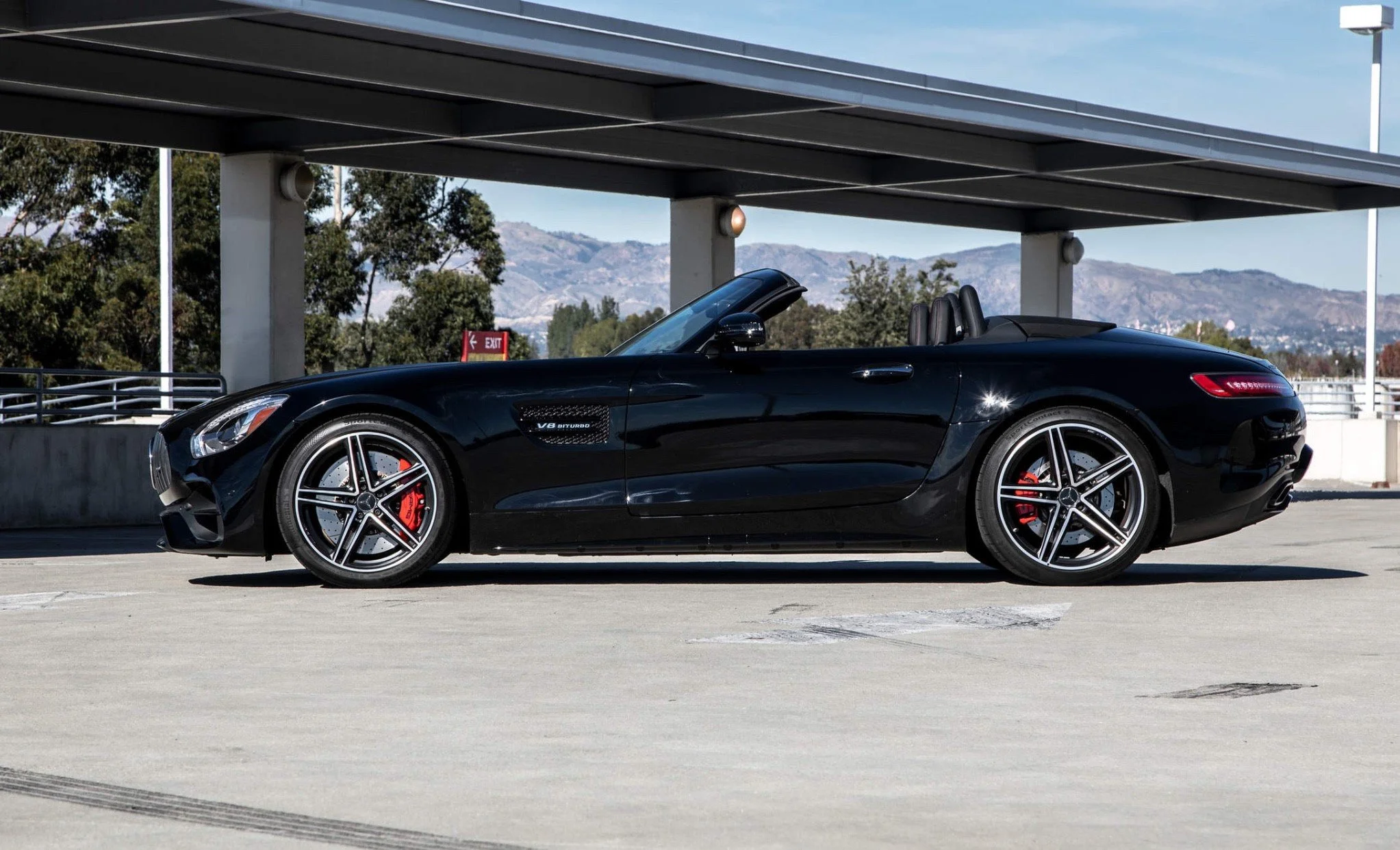 Black convertible sports car parked in a parking lot under a structure with mountain scenery in the background.