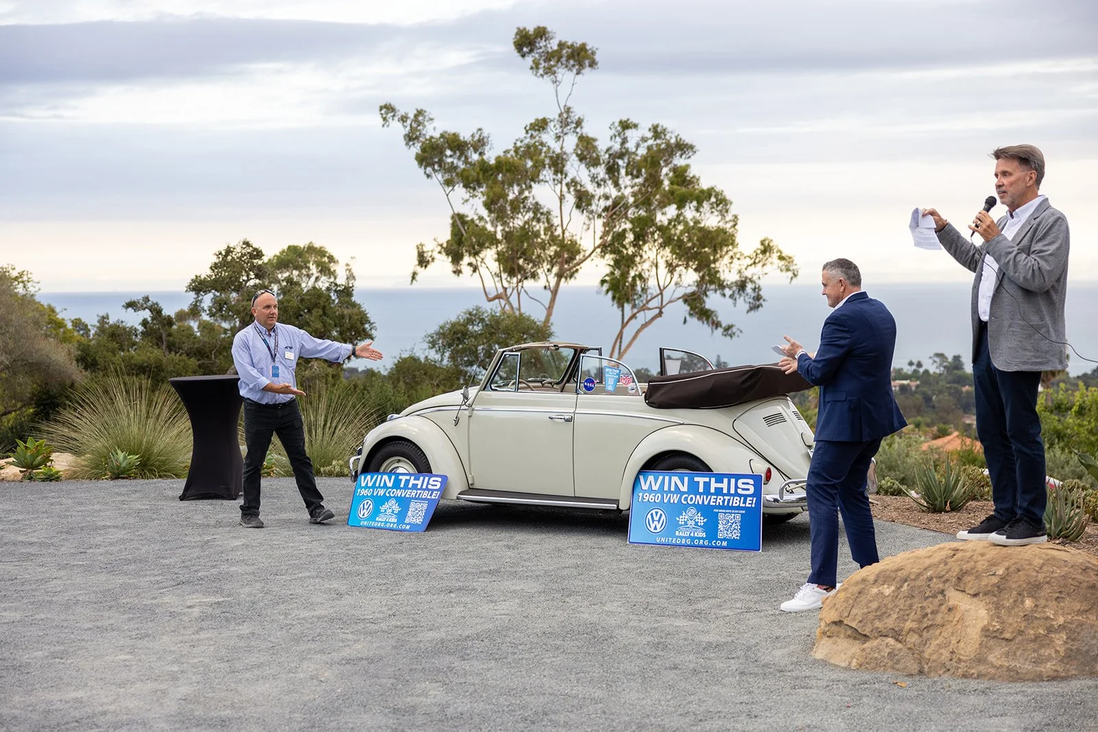 A man in a gray blazer and blue pants is speaking into a microphone, gesturing towards a vintage white VW convertible car. Two other men, one in a suit and the other in a dress shirt, are looking at the car. There are signs on the ground that say 'Wi