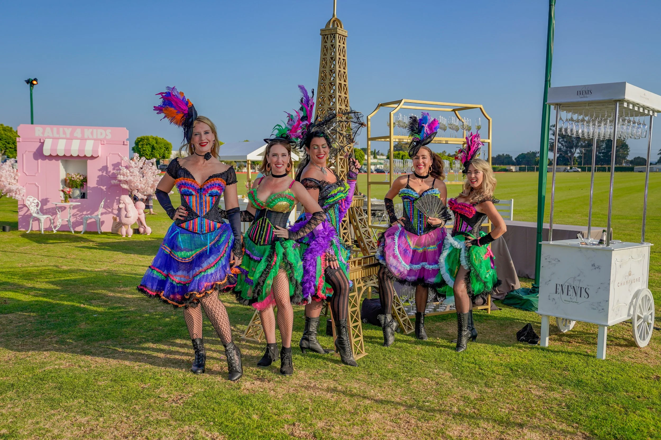 Five women in colorful, vintage-style dresses with feathers and accessories, standing outdoors on a grassy field with a mini Eiffel Tower replica in the background, under a clear blue sky.