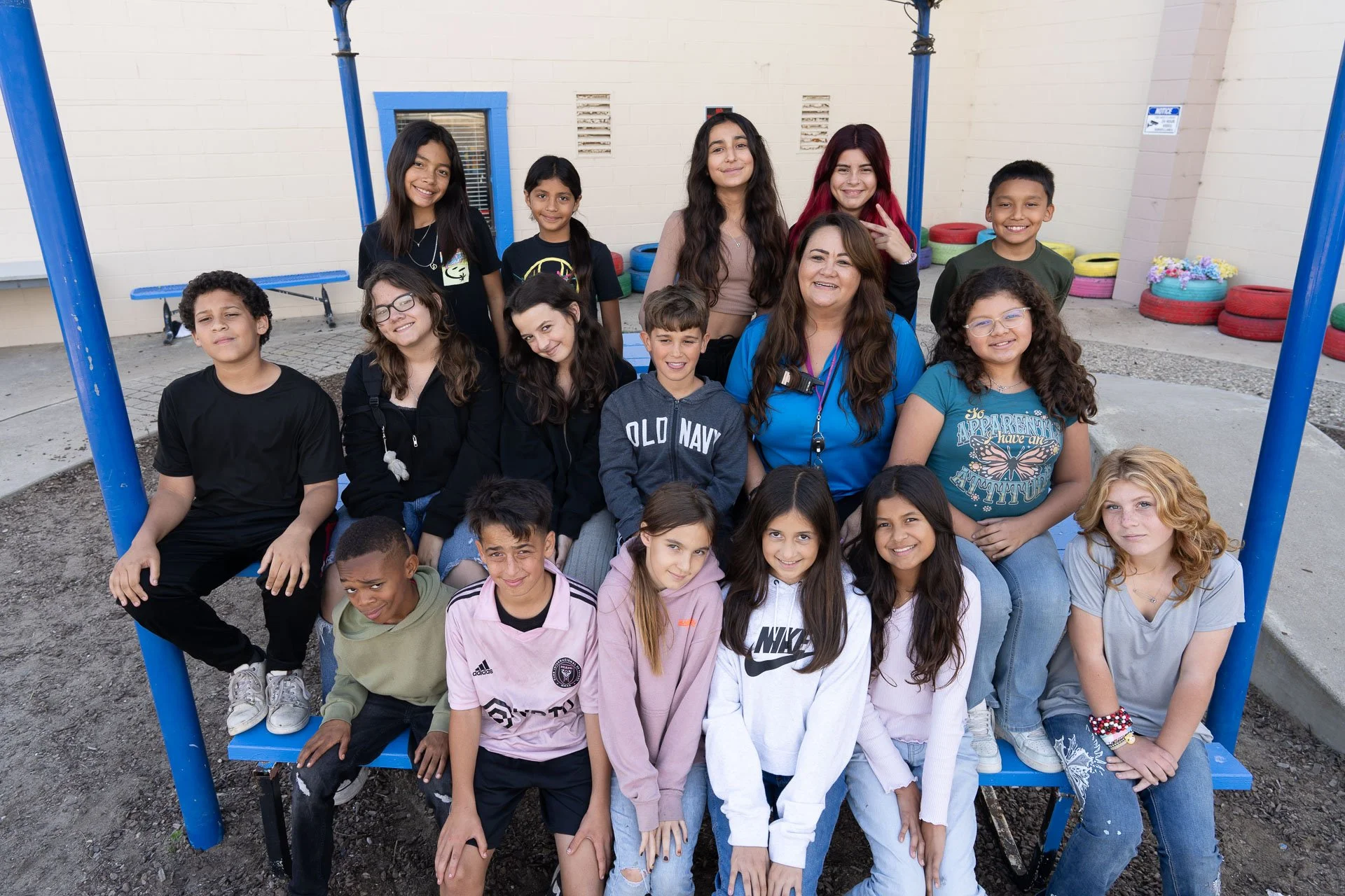 Group of 16 children and a woman, likely a teacher, gathered together on a blue bench outside at a school playground, smiling for the camera.