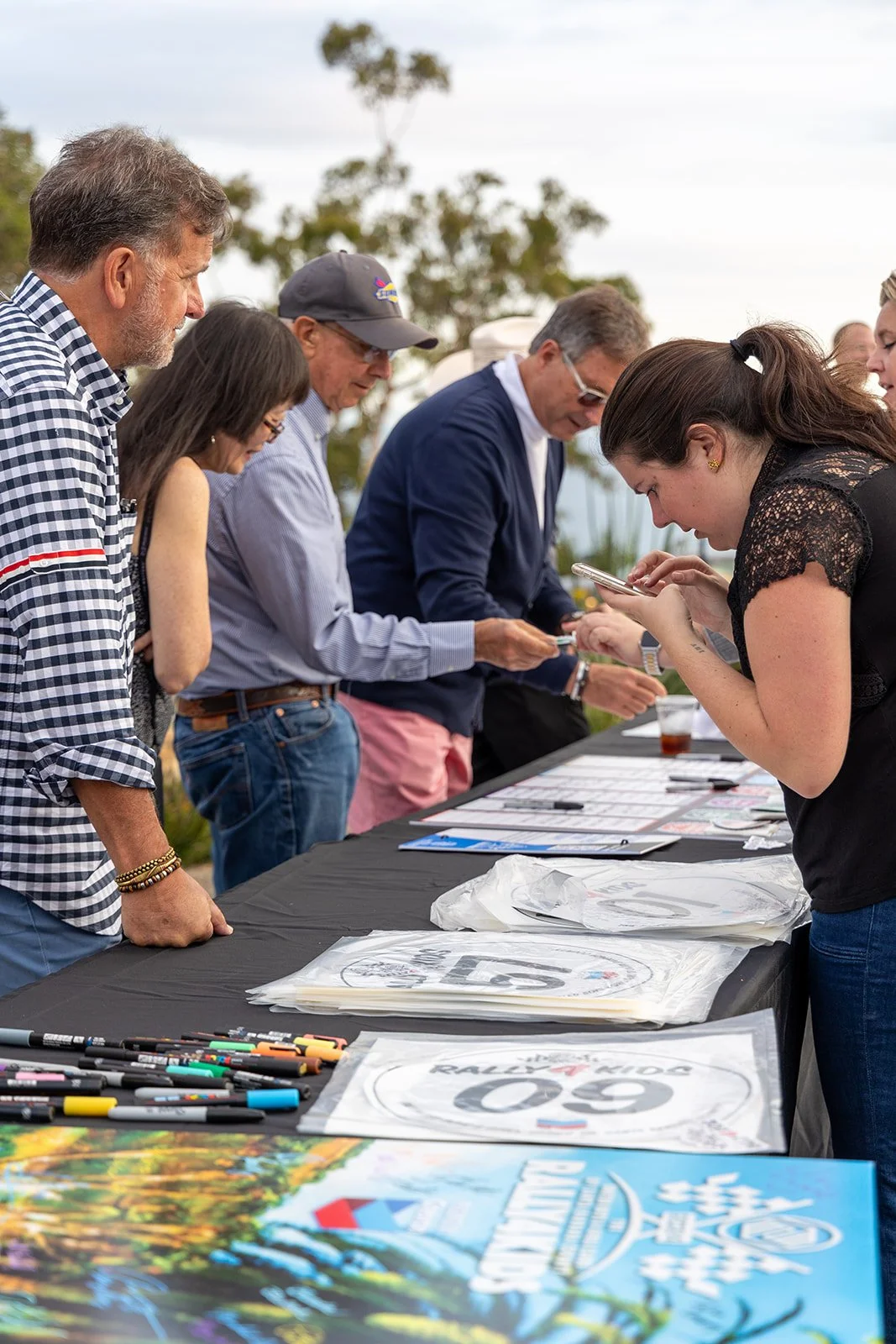 People gathering at a registration table outdoors for an event, signing in and interacting with materials on the table.