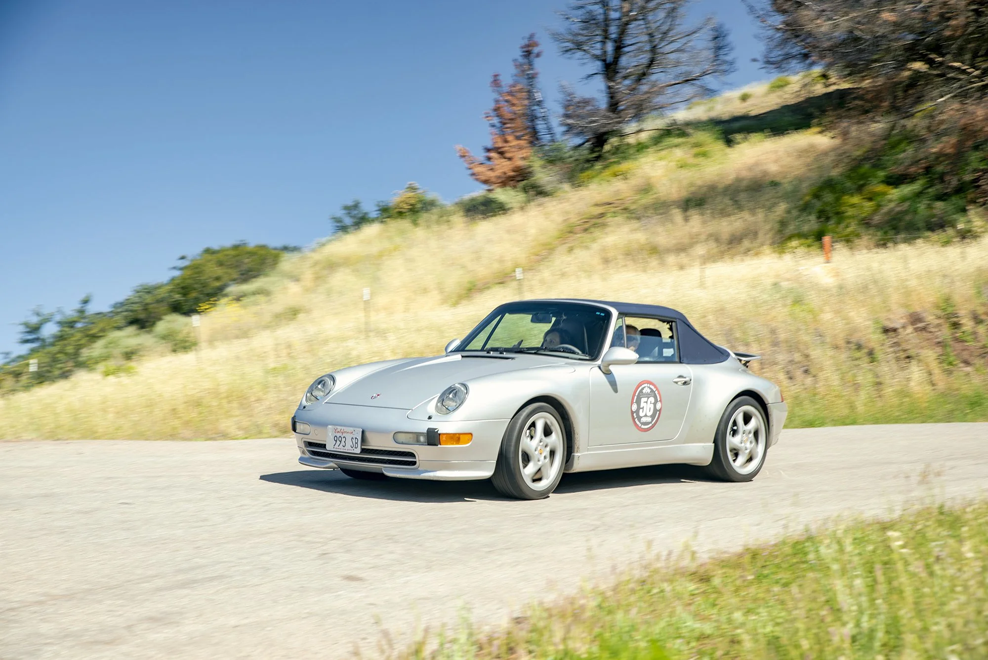 A silver Porsche 911 convertible sports car driving on a winding road with grassy hills and trees in the background.