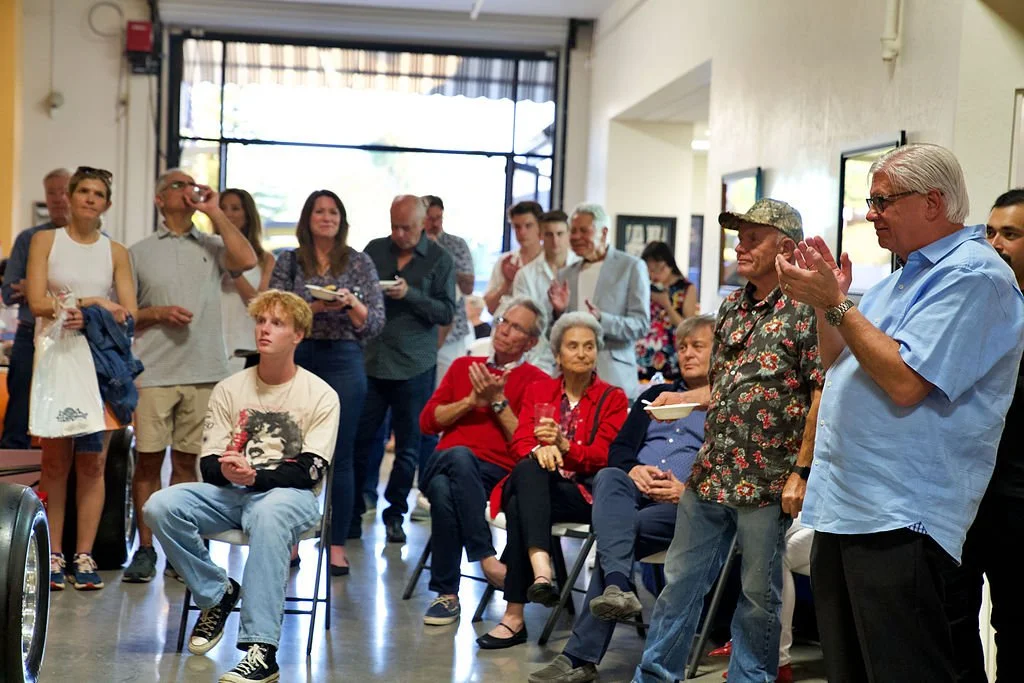 Group of people gathered indoors, some standing and others seated, as an older man claps. Attendees include adults and teenagers, with a woman in red holding a drink and a man with a floral shirt and camouflage cap. The setting appears to be a celebr