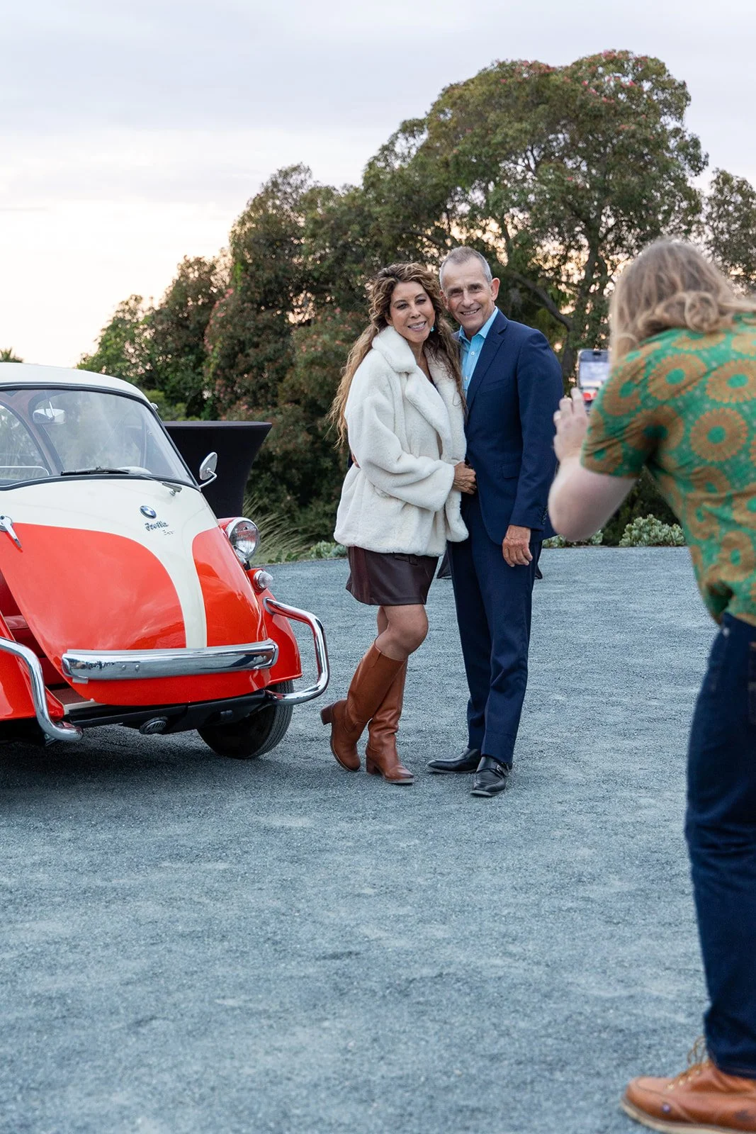A woman in a white coat and brown boots standing next to a man in a blue suit, posing for a photo beside a vintage red and white BMW Isetta on a gravel surface with trees in the background, as another person takes their picture.