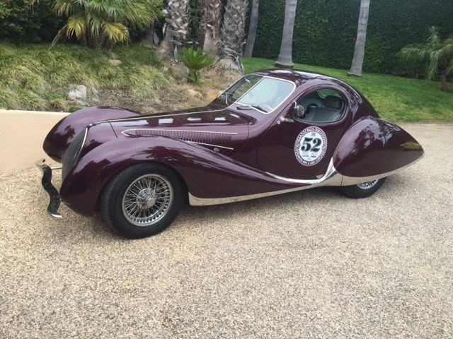 A vintage maroon race car with a racing number 52 on the side, parked on a gravel driveway with trees and bushes in the background.