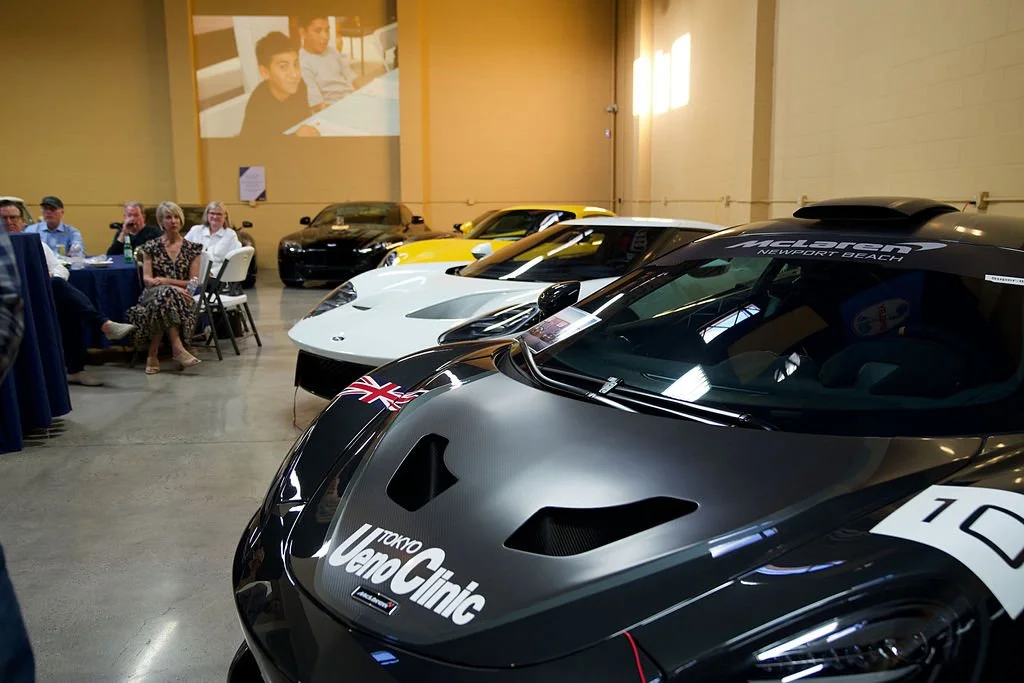 Several high-performance race cars are displayed in an indoor showroom, with a group of people sitting at a table to the left and a large video projection of some individuals on the back wall.