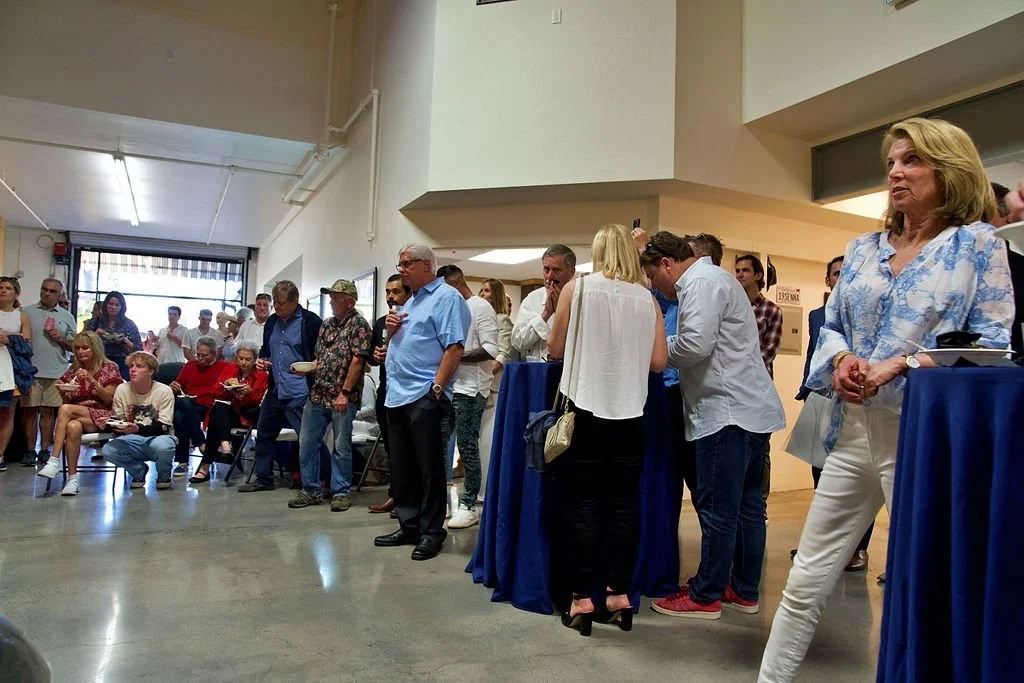 A group of people gathered in an indoor space, some sitting and some standing, with a table covered in a blue cloth where a few people are eating or drinking. The scene appears to be a social or community event.