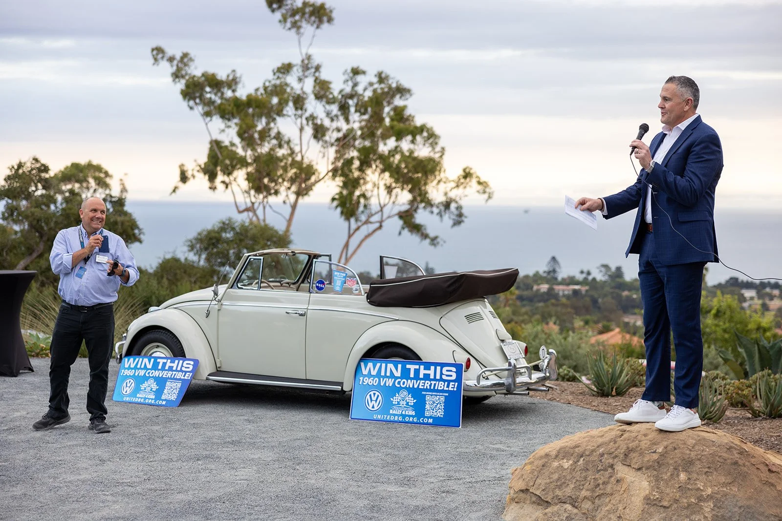 Two men standing outdoors, one holding a microphone and the other a paper, near a vintage white Volkswagen convertible car with signs that read 'WIN THIS 1960 VW CONVERTIBLE!' The background shows trees and a distant landscape, possibly during the da