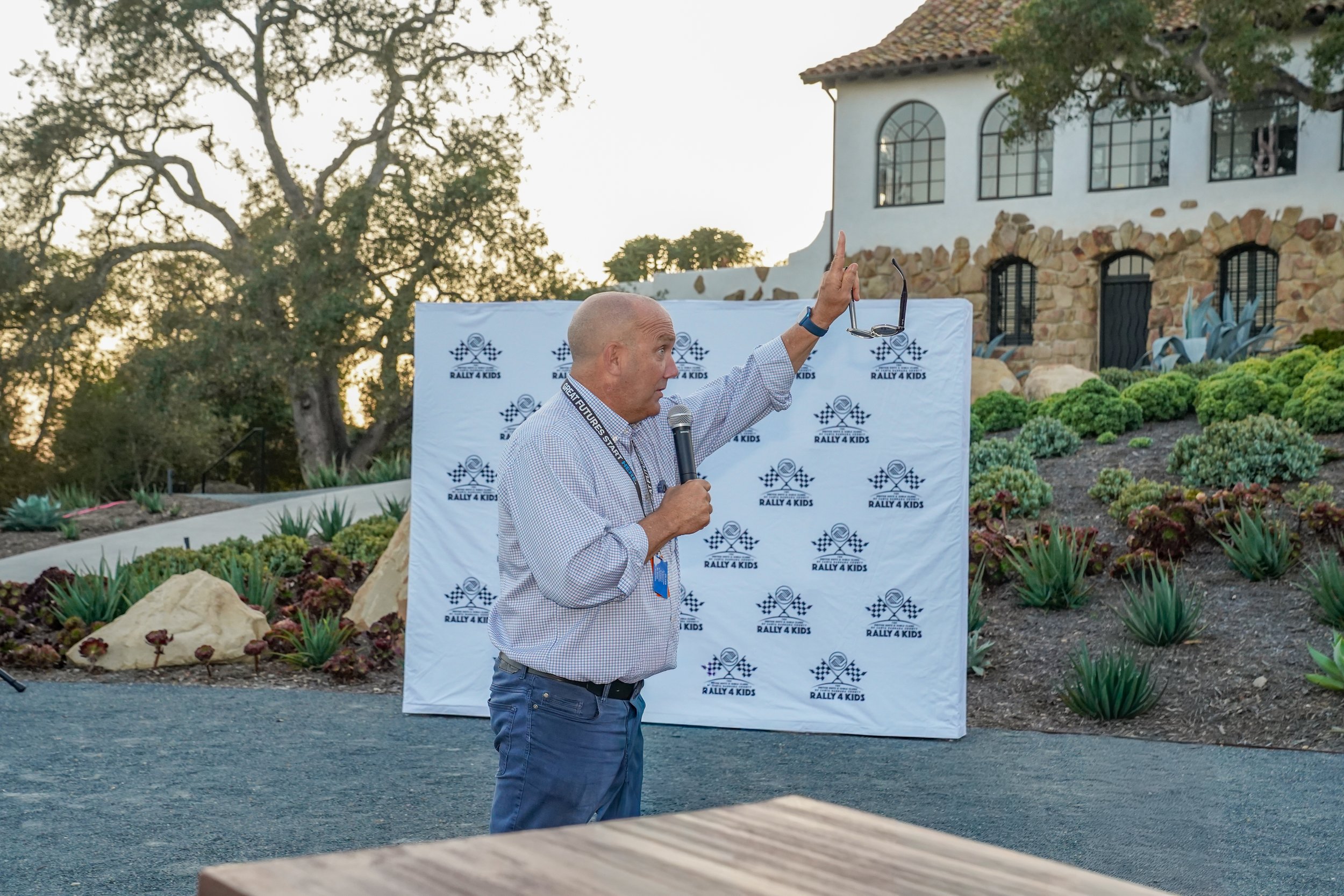 A man with a microphone and lanyard speaking outdoors during sunset, standing in front of a backdrop with 'Rally 4 Kids' logos, with a building and landscaped garden in the background.