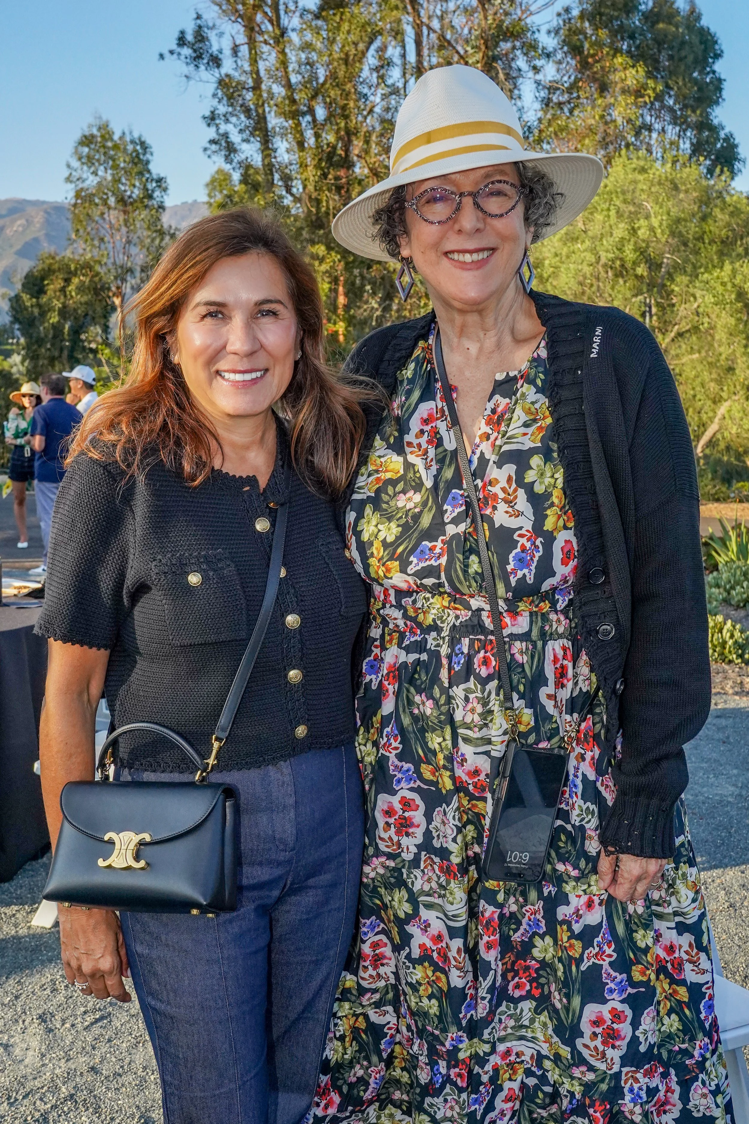 Two smiling women standing outdoors, one wearing a black short-sleeved top, jeans, and a black purse, the other in a floral dress, black cardigan, and wide-brimmed hat with glasses.