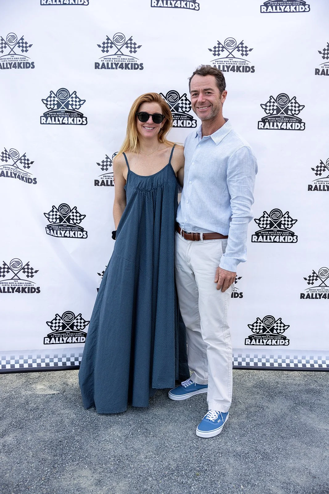 A woman wearing sunglasses and a blue dress standing next to a man in white pants, blue shoes, and a light blue shirt in front of a backdrop with Rally4Kids logo.