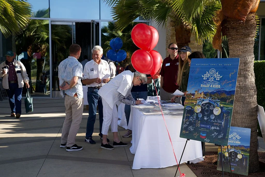 People gather around a registration table at an outdoor event with palm trees, balloons, and a poster for a rally called 'Rally 4 Kids' on April 22, 2022.
