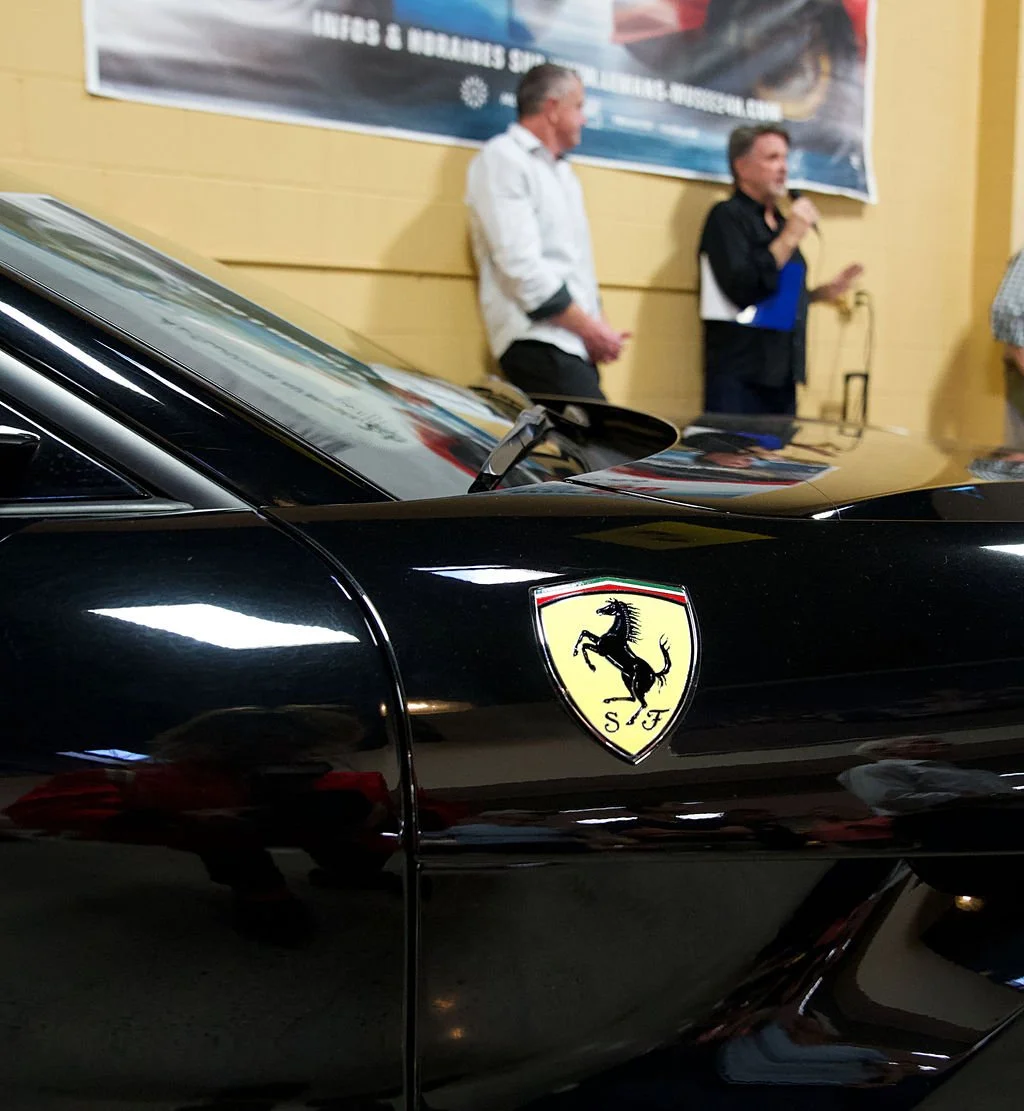 Close-up of a black Ferrari sports car with a yellow shield emblem featuring a black prancing horse, partially showing an event with people and a banner in the background.