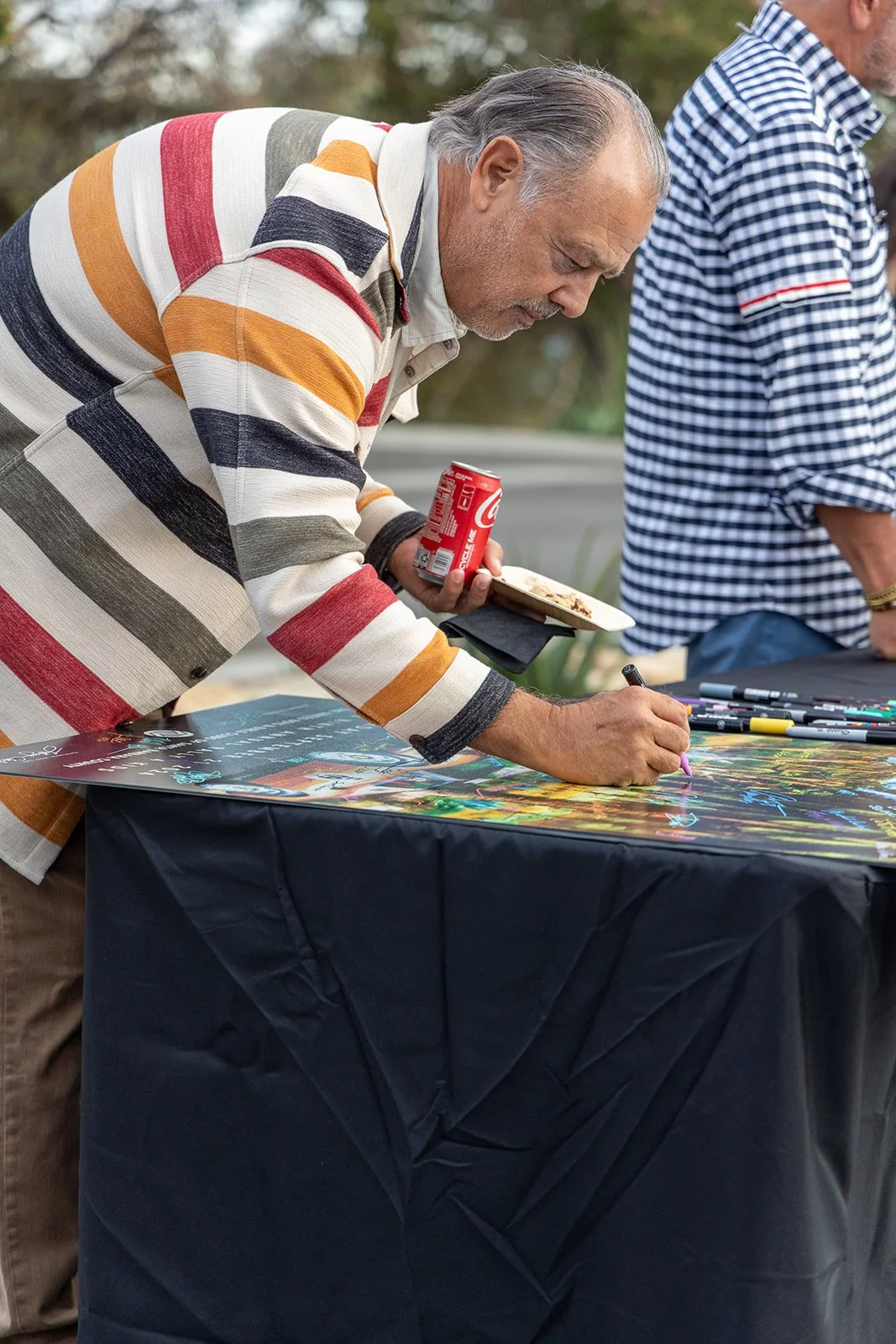 An elderly man in a multicolored striped sweater is signing a poster or artwork on a table, while holding a can of Coca-Cola in his other hand. Another man in a checkered shirt is standing nearby, outdoors.