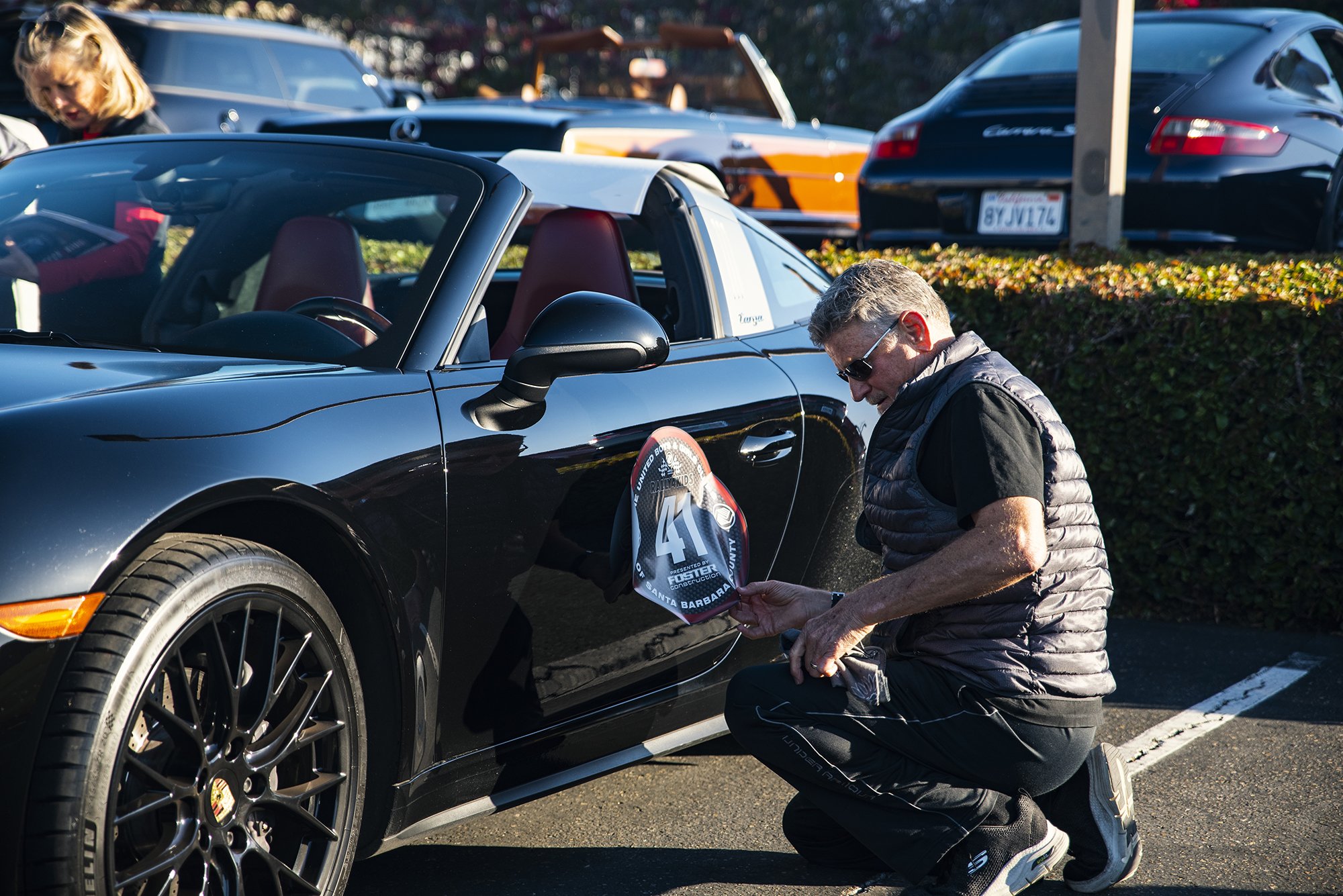 A man kneeling beside a black convertible sports car, attaching a race number sign that says '41' to the side of the car. The scene is in a parking lot with other vehicles and people in the background.