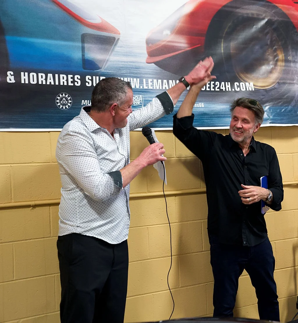 Two men giving each other a high five in front of a race car banner, one holding a microphone and the other holding a notebook, indoors with a yellow brick wall background.