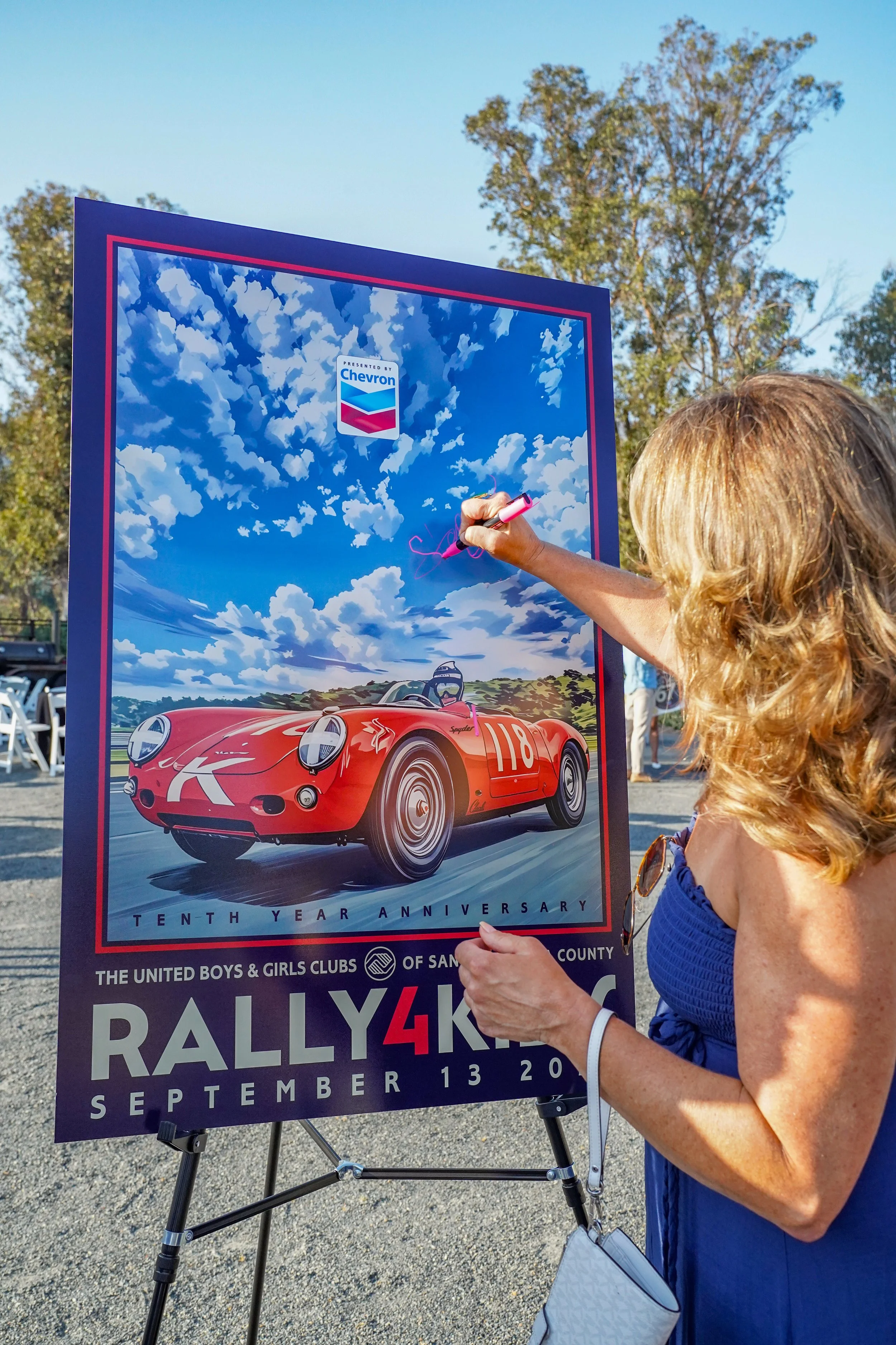 Woman signing a rally event poster featuring a red vintage race car on the day of the event.