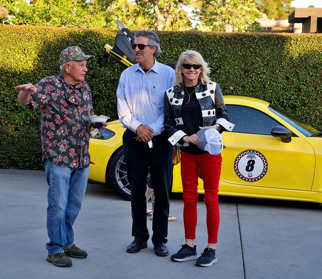 Three people standing outdoors near a yellow sports car, with a man in a floral shirt and camouflage hat, another man in a light blue shirt and sunglasses, and a woman in a black and white jacket with red pants, all smiling and engaged in conversatio
