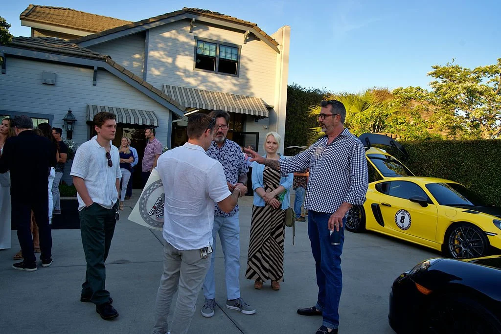 Group of people socializing outdoors in front of a white house with a yellow sports car and a black sports car parked nearby.